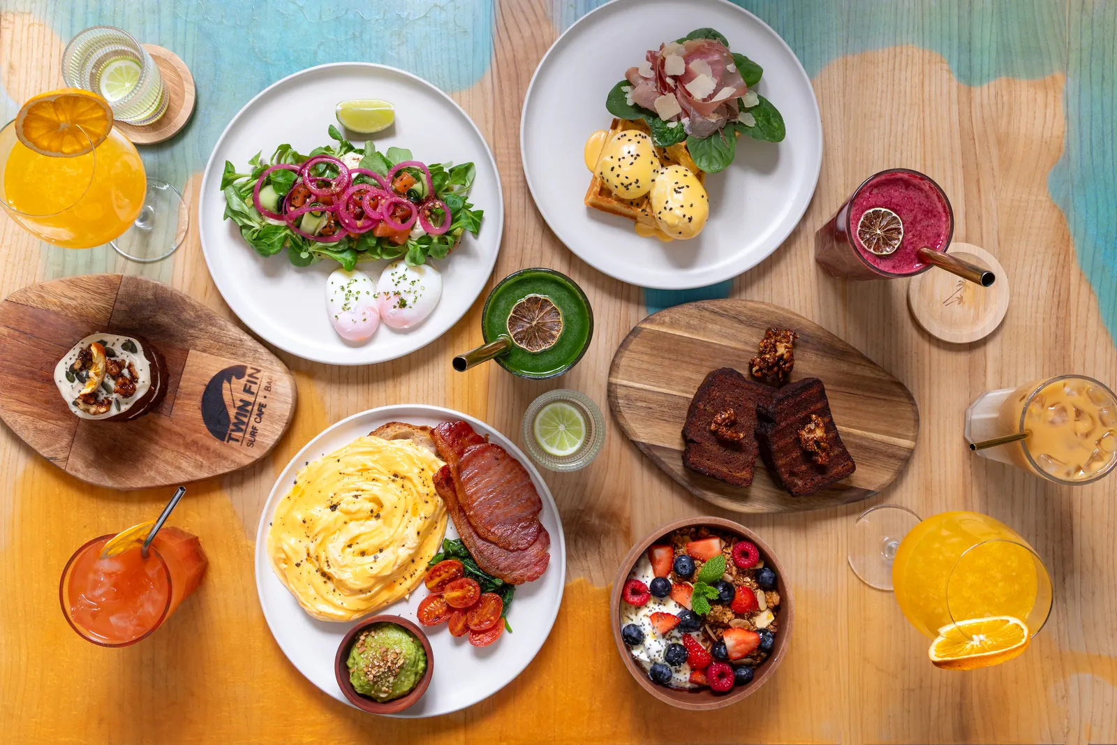 Overhead view of a Twin Fin table with salmon sourdough, a benedict waffle, scrambled eggs with bacon, banana bread, a granola bowl, smoothies, orange juice and mimosas